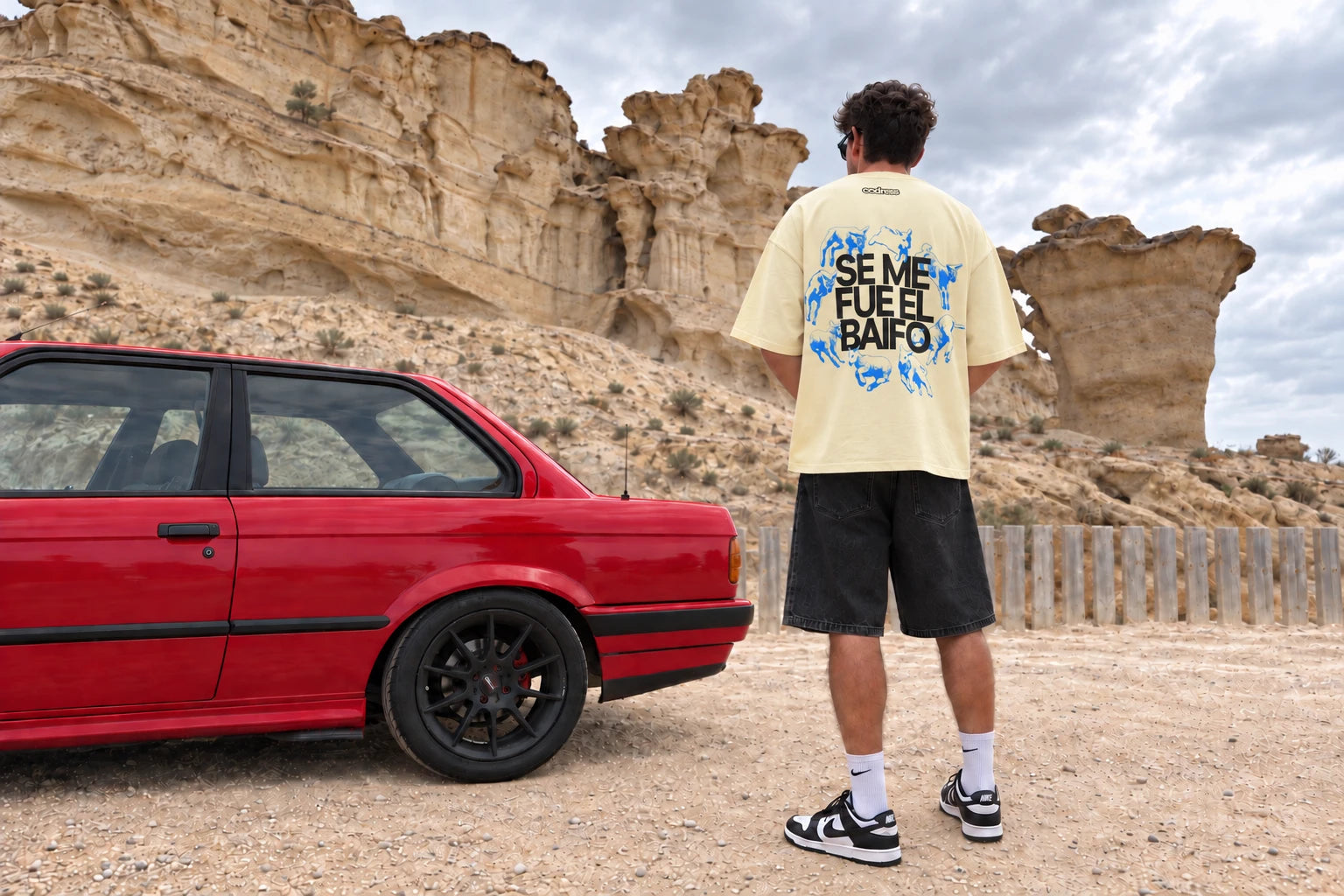Modelo de espaldas con la camiseta amarilla Baifo junto a un coche rojo y un paisaje de roca.